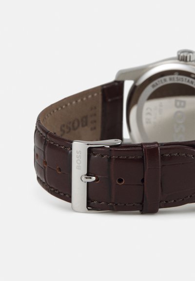 Bracelet de montre en cuir marron avec motif crocodile, équipé d'une boucle en argent portant le nom de la marque "BOSS". Boîtier en acier inoxydable avec résistance à l'eau.