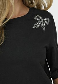 Tricou negru din bumbac cu guler rotund, având un design decorativ cu fundă strălucitoare pe umărul stâng, realizat din pietricele strălucitoare.