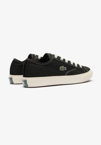 Sorte lerretsko med hvite snører, gummisåler og en grønn Lacoste krokodillelogo på siden. Klassisk design med lav profil.