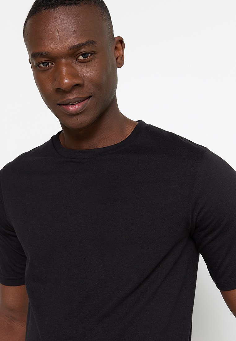 Un jeune homme aux cheveux courts portant une chemise noire ajustée à manches courtes, souriant doucement sur un fond blanc uni.