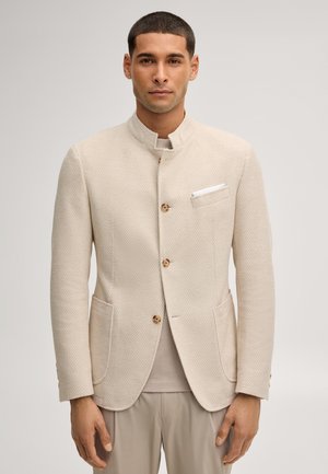 Mann trägt eine helle beige strukturierte Jacke mit Stehkragen und Holzknöpfen, kombiniert mit neutralen Hosen und hellem Hemd.