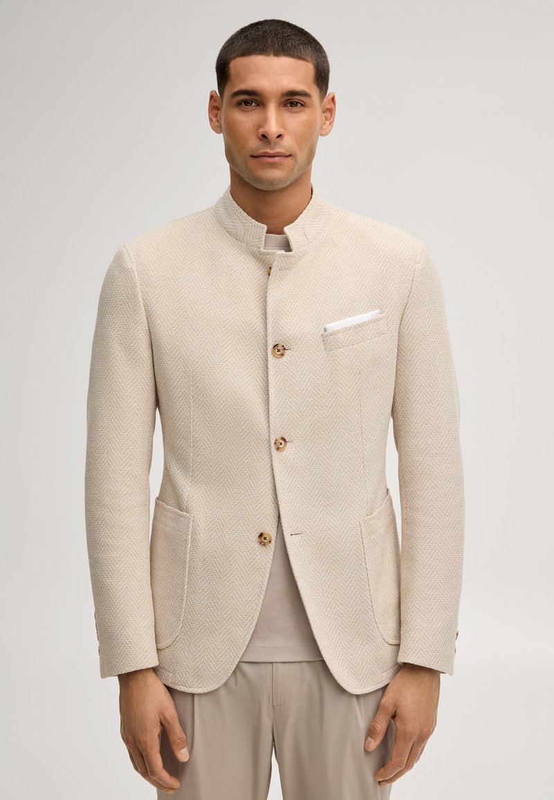 Mężczyzna noszący jasnobeżową, teksturowaną kurtkę z kołnierzem typu stójka i drewnianymi guzikami, w połączeniu z neutralnymi spodniami i jasną koszulą.