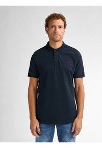 Donkerblauwe polo-shirt van katoen met een kraag, een twee-knopengulp en een klein rood logo op de borst, gecombineerd met denim jeans.