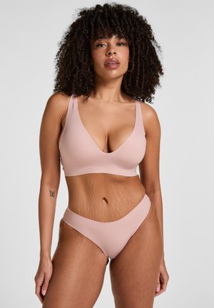 Frau mit lockigem Haar, die einen hellrosa Bralette und passende Unterwäsche trägt, steht vor einem schlichten weißen Hintergrund mit neutralem Ausdruck.