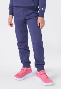 Kind in paarse sweatpants en een bijpassende sweatshirt met felroze sportschoenen met witte zolen op een witte achtergrond.