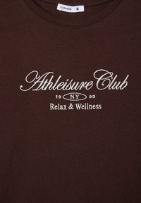 Brązowa bawełniana koszulka z okrągłym dekoltem. Zawiera białe, haftowane napisy "Athleisure Club", "NY", "Relax & Wellness".