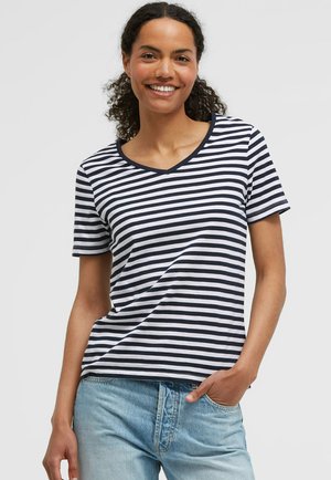 Lächelnde Frau mit lockigem Haar, die ein schwarz-weiß gestreiftes V-Neck-T-Shirt und hellblaue Jeans trägt, mit der rechten Hand in der Tasche.