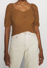 Brun, texturerad blus med puffärmar och fyrkantig halsringning. Har broderade detaljer och en åtsittande siluett. Matchas med vita jeans.