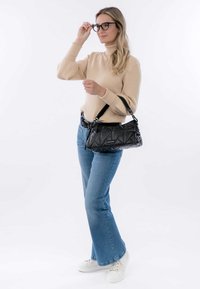 Zwarte gewatteerde handtas met een schouderband, vastgehouden door een persoon in een beige ribbentrui en blauwe flared jeans, staand tegen een neutrale achtergrond.