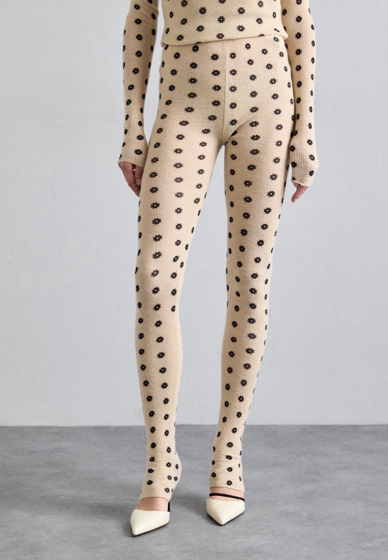 Leggings beige a coste con un motivo floreale nero, abbinati a tacchi bianchi a punta. Presentano maniche lunghe e una linea aderente.