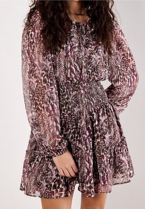 Femme portant une robe à manches longues, longueur genou, avec un imprimé animalier mixte dans des nuances de violet, rose et blanc, main posée sur la jupe.