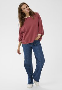 Mosolygó nő, lazán illeszkedő mauve színű pulóverben, fehér díszítéssel, kék farmerben, fehér cipőben, az egyik kezét zsebébe dugva áll egy fehér háttér előtt.