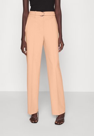 Frau trägt beige weite High-Waist-Hosen mit einem Ringgürtel und schwarzen Riemchensandaletten mit Absatz vor einem einfarbigen hellen Hintergrund.
