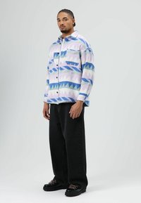 Meerpaars flanellen overhemd met geometrische patronen in blauw, groen en paars, met een voorkantzak en zwarte knopen, gedragen met een donkere jeans.