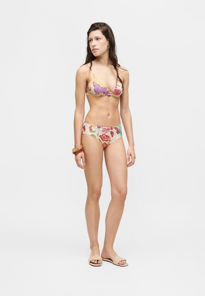 Femme debout portant un bikini à imprimé floral, des sandales beiges et un bracelet en ambre, sur un fond clair uni.