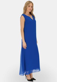 Niebieska sukienka maxi z dekoltem w szpic i krótkimi rękawkami. Posiada ozdobny srebrny dekolt oraz lekką, przezroczystą tkaninę o zwiewnej fakturze. Czarnie szpilki dopełniają stylizację.