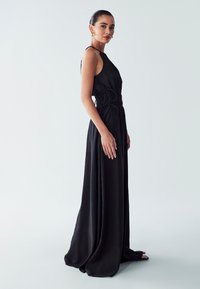 Black sleeveless dress with a cinched waist and flowing skirt. Made from smooth fabric with a slight sheen, featuring intricate tie details.