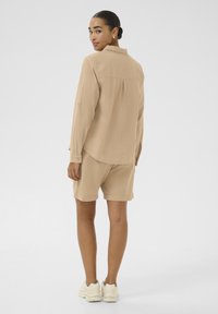 Beige overhemd met lange mouwen, gedragen met bijpassende shorts. Heeft een relaxte pasvorm en frontzakken, gecombineerd met lichtgekleurde sneakers.