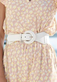 Breed witleren riem met een ronde gesp, gedragen over een lichtroze jurk met een bloemenpatroon in wit en geel.