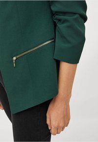 Grön blazer med en strukturerad passform, med en ficka med dragkedja på sidan och upprullad manschettdetalj. Slät tygtextur.