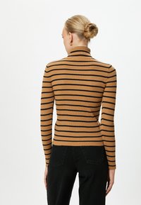 Gestreifter braun-schwarzer, ribbeliger Rollkragenpullover mit langen Ärmeln, körperbetontem Design und einfarbigem Rücken. Getragen mit schwarzen Hosen.