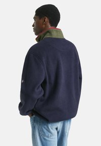 Marineblaue Fleece-Pullover mit grünem Innenkragen und roten Akzenten. Verfügt über eine lockere Passform und gerippte Bündchen, die einen strukturierten Stoff zeigen.