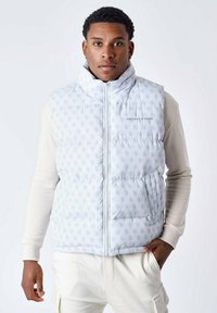 Gilet imbottito grigio chiaro con motivo logo su tutta la superficie, chiusura con zip, colletto alto e tasche laterali. Indossato sopra una maglietta a maniche lunghe color crema.