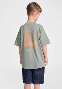 Grön bomullströja med en stor orange grafisk design på baksidan, som visar texten "the stadium ANTHENS" och en karaktärsdesign. Korta ärmar.