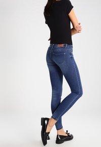 Vaqueros de denim azul ajustados con un acabado desgastado, que cuentan con bolsillos traseros y una etiqueta de cuero en la cintura, combinados con bailarinas negras.