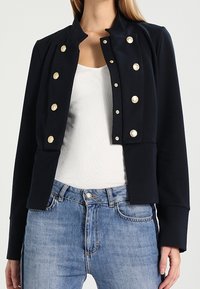 Marinblå croppad jacka med guldknappar, strukturerad design och mjukt tyg; kombinerad med en vit topp och ljusblå jeans.