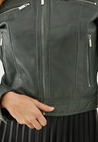 Tummanvihreä nahkatakki, jossa on metalliset vetoketjut, muotoiltu malli ja vetoketjulliset taskut. Sujuva pinta ja näkyvät ompelutiedot.