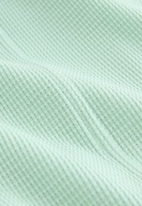 Texturovaná látka v mátově zelené barvě s jemným mřížkovým vzorem. Lehká a jemná, vykazuje hladký, rovnoměrný povrch.
