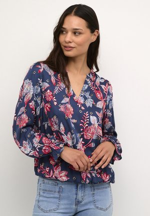 Blusa navy con intricati motivi floreali rossi e bianchi, con scollatura a V e maniche arricciate, abbinata a jeans azzurri chiari.