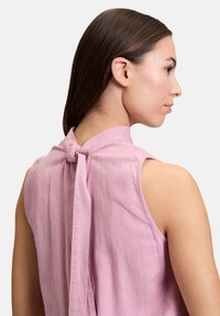 Roze mouwloze blouse met een hoge halslijn en een strikdetail op de rug. Gemaakt van zachte, lichte stof met een gladde textuur.