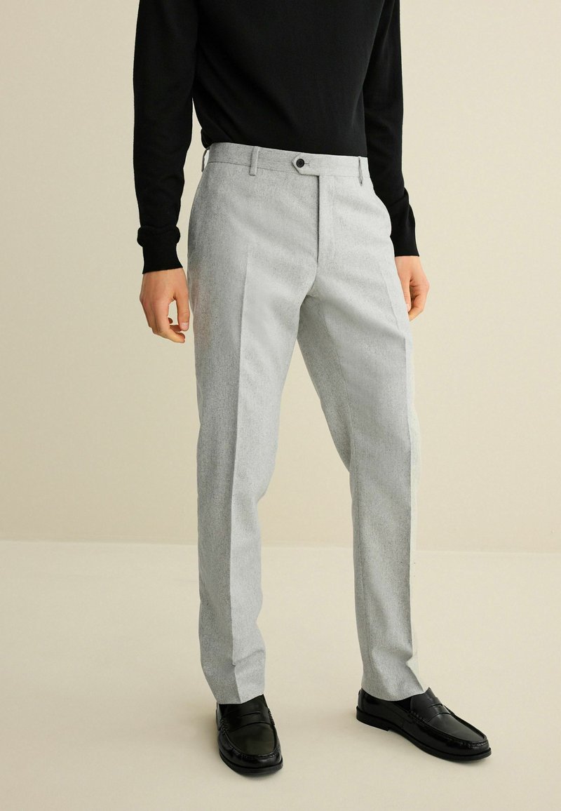Hellgraue Hose mit strukturiertem Finish, mit flacher Vorderseite, seitlichen Taschen und Knopfverschluss, getragen mit schwarzen Schuhen und einem schwarzen Pullover.