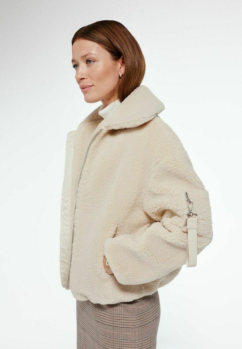 Kremowa kurtka z sztucznego futra o szerokim kołnierzu, w oversize'owym kroju, z boczną kieszenią. Posiada pasek z klipsem na rękawie. Teksturowana powierzchnia.