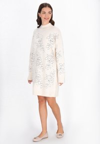 Rochie tricotată crem cu un guler înalt, având modele brodate cu frunze negre. Mâneci lungi și o croială relaxată. Asortată cu balerini bej.