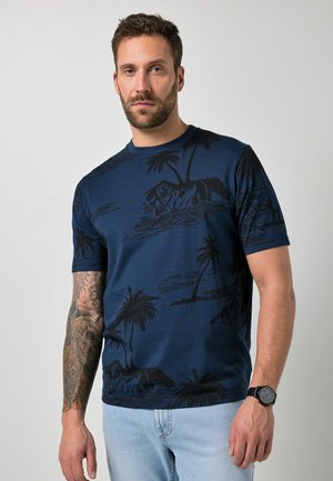 Mężczyzna z brodą, ubrany w niebieską koszulkę z tropikalnym motywem i jasne dżinsy, pokazujący wytatuowane prawe ramię i czarny zegarek na rękę.