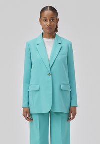 Turquoise blazer met een enkele knoopsluiting, twee voorzakken en een geknipt kraag; gecombineerd met bijpassende broek. Glad stoftextuur.