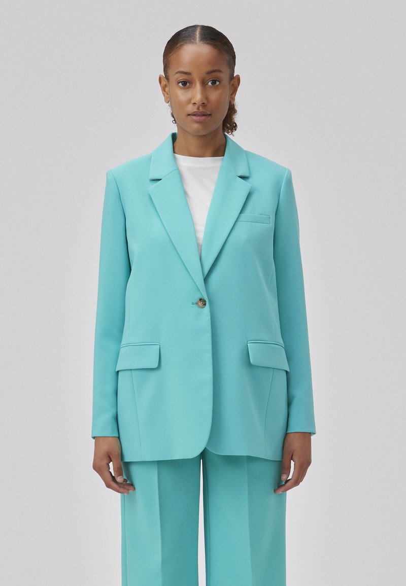Turquoise blazer met een enkele knoopsluiting, twee voorzakken en een geknipt kraag; gecombineerd met bijpassende broek. Glad stoftextuur.