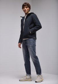 Veste à capuche navy avec fermeture éclair et deux poches avant, associée à un jean bleu décoloré et des sneakers beiges, sur fond neutre.