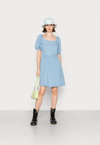 Vestido de ganga azul claro com mangas curtas em formato de balão e decote quadrado, combinado com um chapéu de balde em tons pastéis e botas pretas com atacadores.