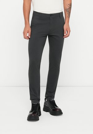 Slim-fit dark gray trousers made of smooth fabric, featuring a button closure, side pockets, and a tapered leg design. Worn with black boots.
