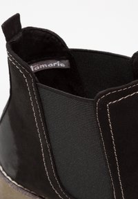 Gros plan sur une botte noire à cheville avec panneau latéral élastique, coutures blanches contrastantes, languette de tirage et étiquette de marque en tissu à l'intérieur.