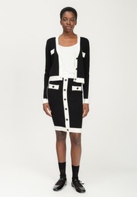 Schwarz-weißer Cardigan und Rock, mit Knopfdetails und kontrastierenden Streifen, aus strukturiertem Stoff gefertigt. Schwarze Schuhe und Socken ergänzen das Outfit.