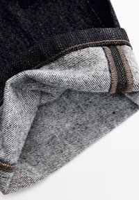 Tessuto denim in sfumature di grigio con una superficie leggermente strutturata, caratterizzato da un polsino blu scuro e dettagli di cucitura marrone a contrasto.