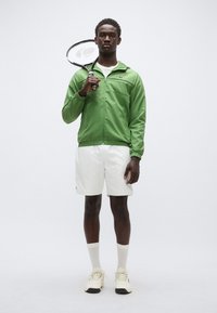 Zelena najlonska jakna z zadrgo spredaj, bela majica, bele kratke hlače in teniški lopar. Model nosi bež superge in bele nogavice.