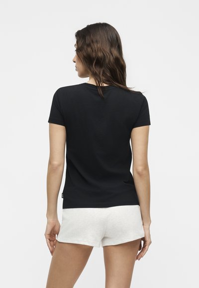 Γυναίκα με κυματιστά καστανά μαλλιά φοράει ένα απλό μαύρο κοντομάνικο μπλουζάκι και λευκά σορτς, στέκεται με την πλάτη προς το λευκό φόντο.