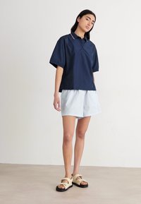 Polo shirt blu navale a maniche corte con colletto e accento rosso, abbinato a pantaloni corti a righe chiare. Il modello indossa sandali beige. Sfondo semplice.