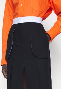 Mustat hameet, joissa on suuret, pyöreät taskut, valkoinen vyötärö ja sivuhalkio, yhdistetty kirkkaan oranssiin napitettava paitaan.